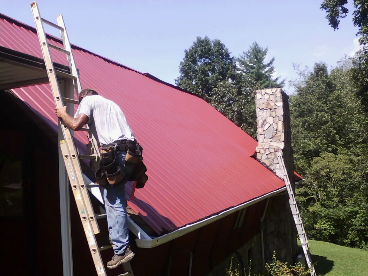 Roofer on a ladder beside a red metal roof and stone chimney — Soffit & Fascia Repair by Roofing Brothers in Cle Elum